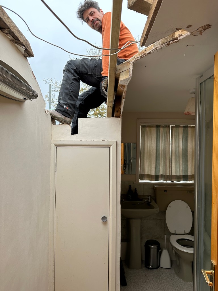 An open section of roof, revealing the bathroom below. A roofer sits on part of the timber structure supporting the roof.